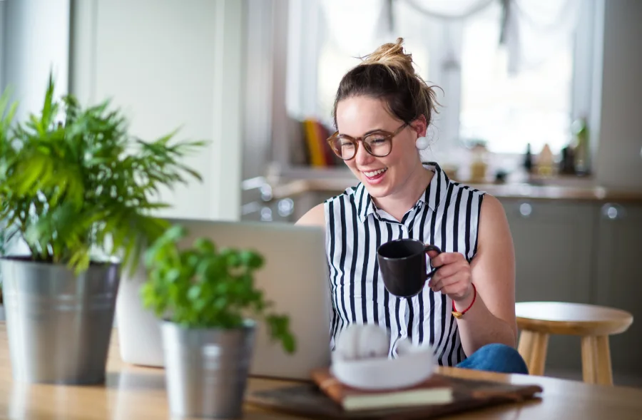 Boldogan nevető, szemüveges, fiatal nő a laptopja előtt ül és dolgozik, kezében bögrével a saját konyhájában.